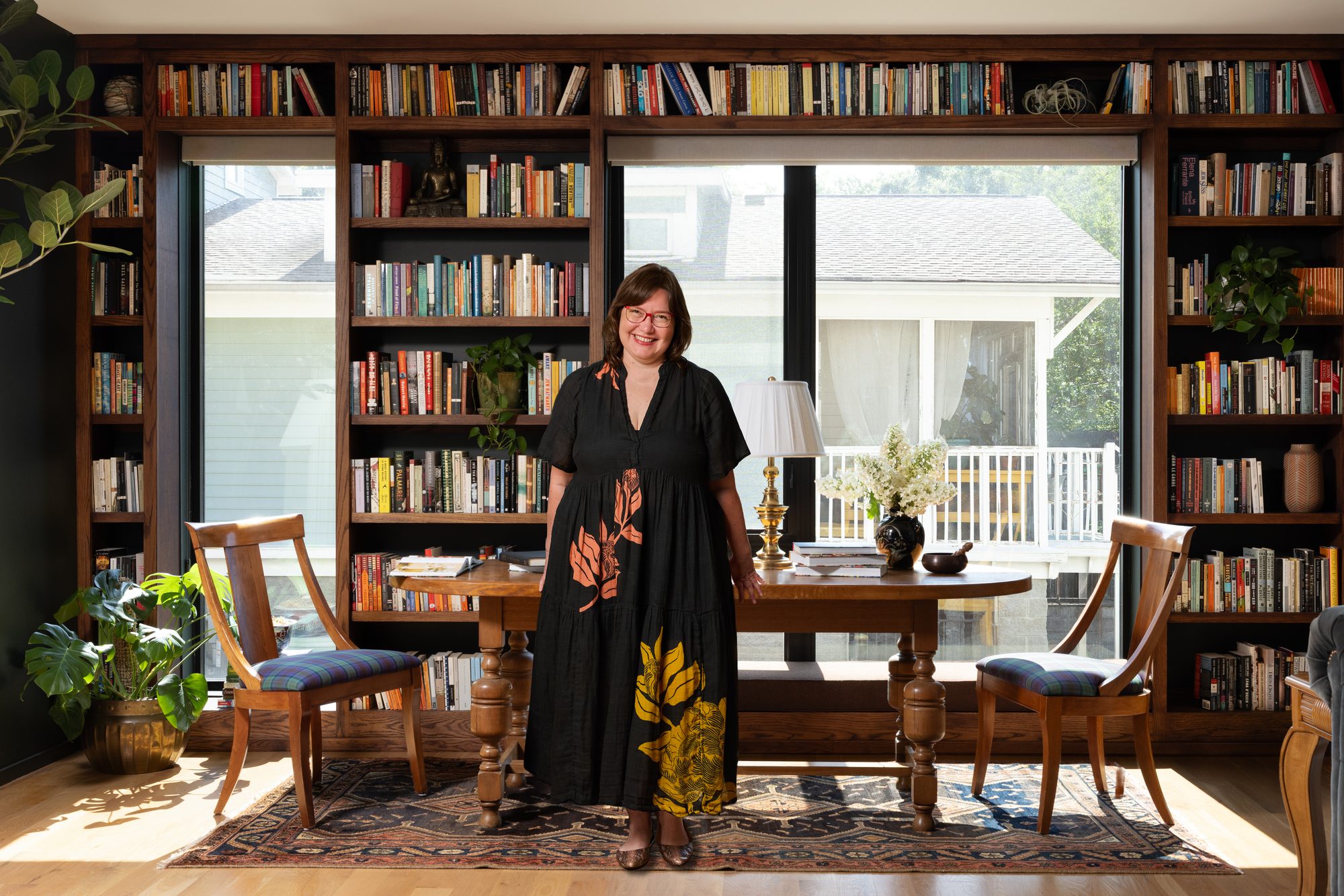Kristin Keyes standing at an antique writing table before a wall of built-in bookshelves in her East Nashville library, wearing a black dress with orange and gold botanical print