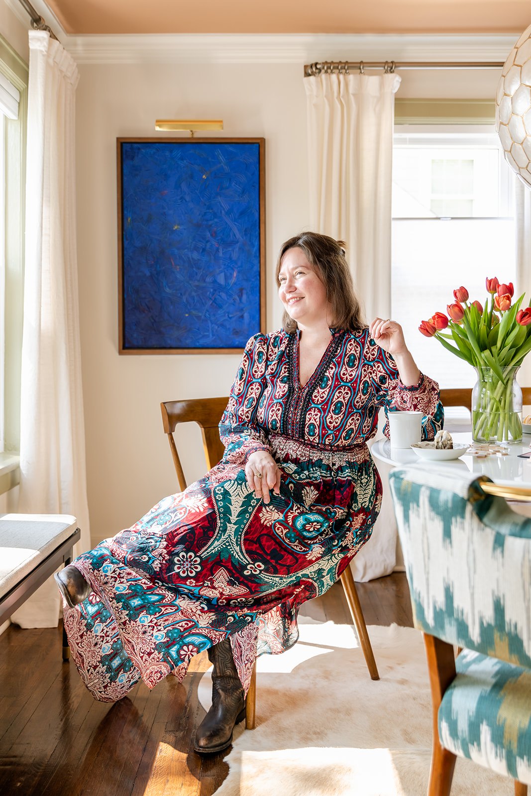 Kristin Wiens Keyes, founder of Kristin Keyes Interiors, seated in a midcentury chair in the sunlit dining nook, in a patterned dress and cowboy boots, beside a cobalt-blue painting and a vase of red tulips