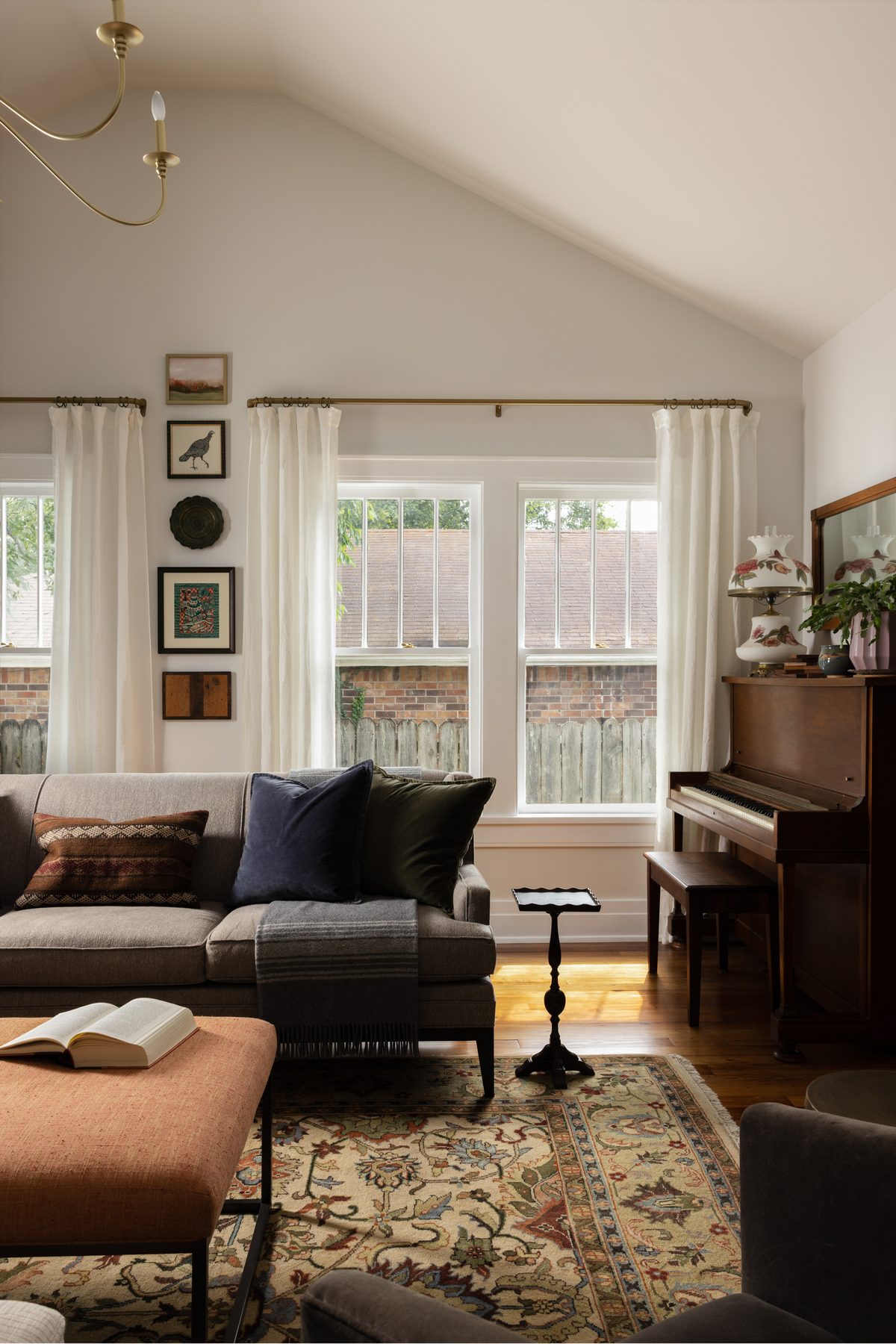 A sofa with velvet pillows beside the family's upright piano, under a vaulted ceiling with a gallery wall and brass curtain rods