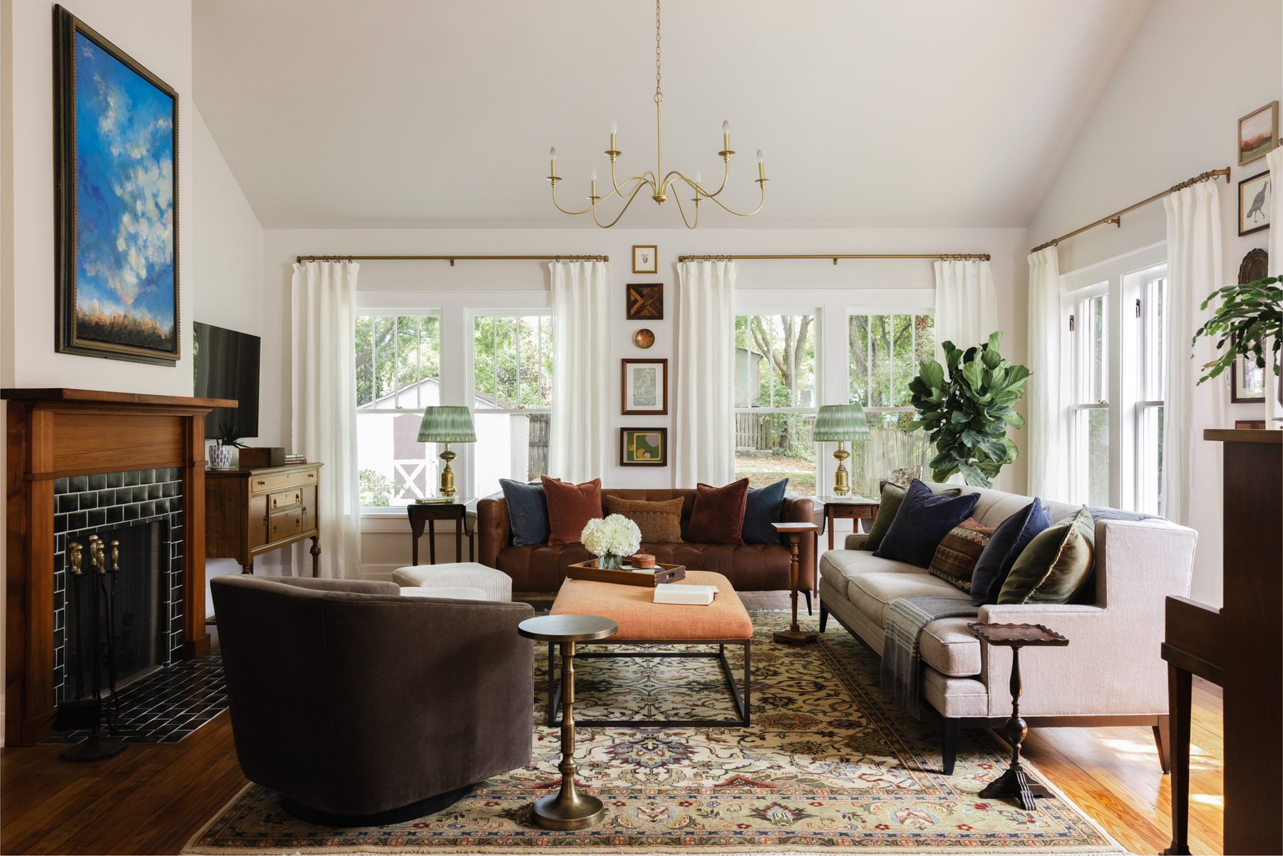 A vaulted great room with two facing sofas, an orange linen ottoman, an upright piano, a dark-green-tiled fireplace with a large blue landscape painting, and a brass chandelier