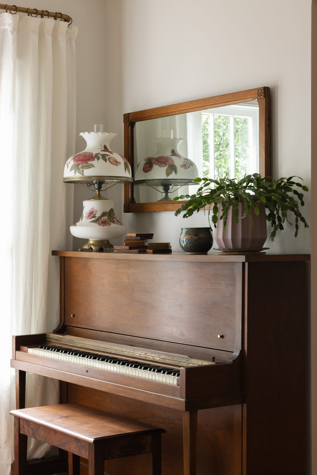 The Great Room's upright piano styled with a floral hurricane lamp, a wood-framed mirror, and a potted plant