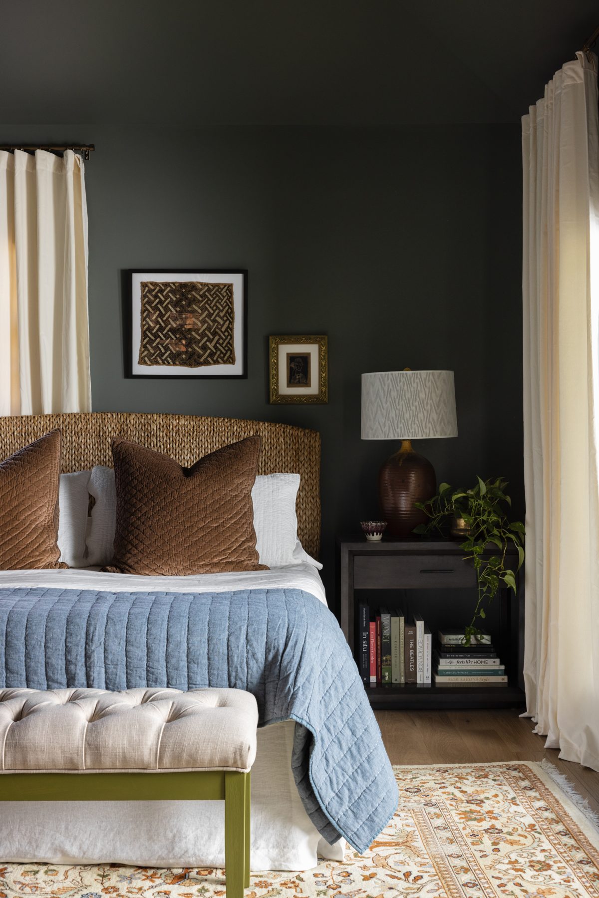 A deep green bedroom corner: a woven seagrass headboard with brown velvet pillows, a blue quilt, a pottery lamp on a dark nightstand stacked with books, and a pothos trailing beside cream drapery
