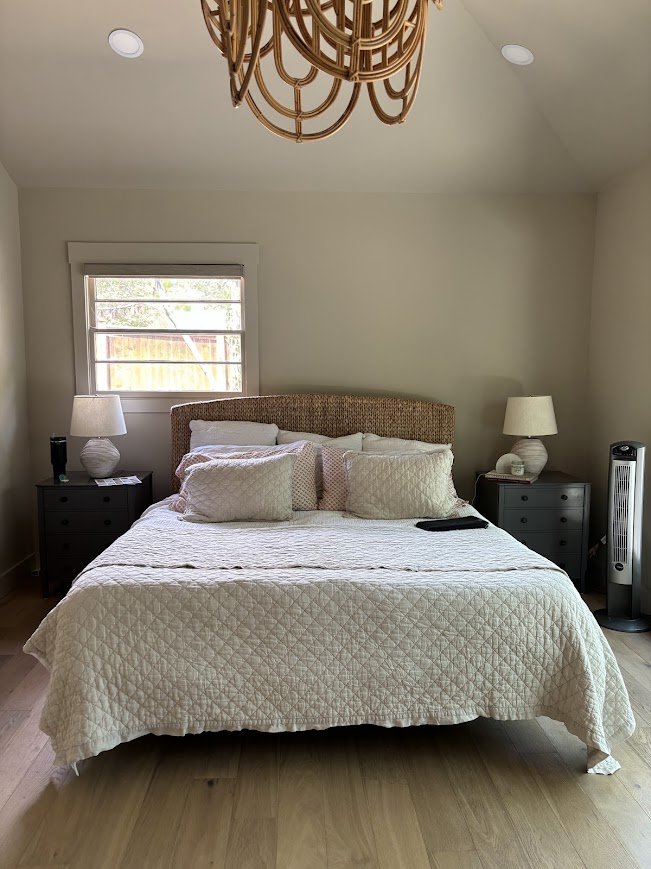 The bedroom before: beige walls, the same seagrass headboard, a tan quilt, mismatched dark nightstands, and a tower fan in the corner
