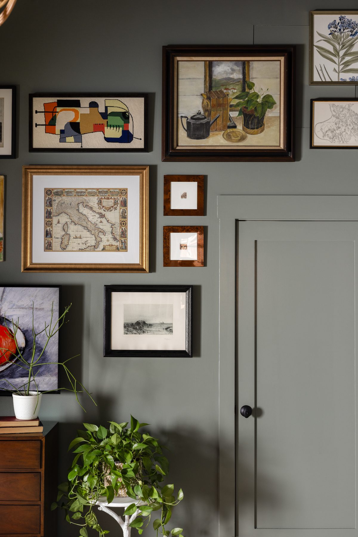 A gallery wall of vintage paintings, an antique map of Italy, a needlepoint abstract, and small burl-framed prints wrapping around a door painted the same green as the walls