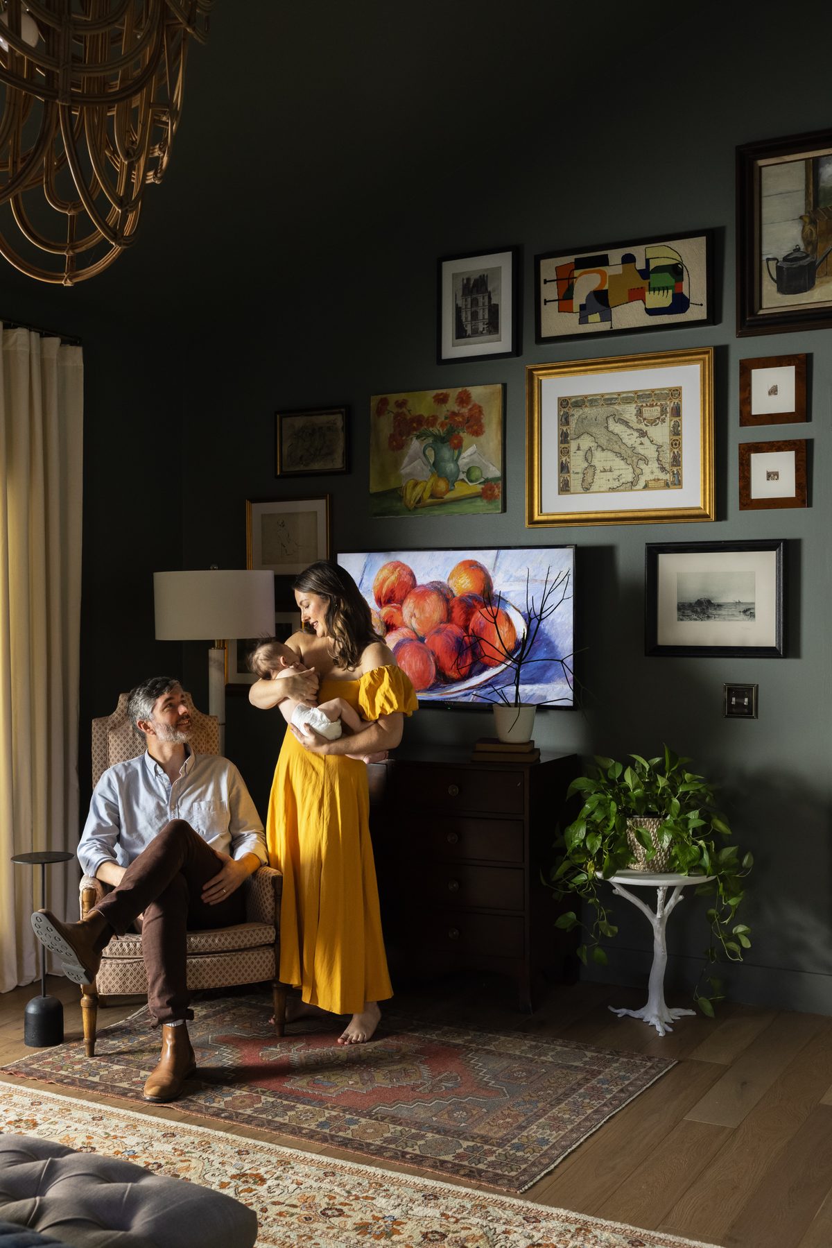 A couple in their finished green bedroom beneath a rattan chandelier, the mother in a yellow dress holding their baby, beside the vintage gallery wall