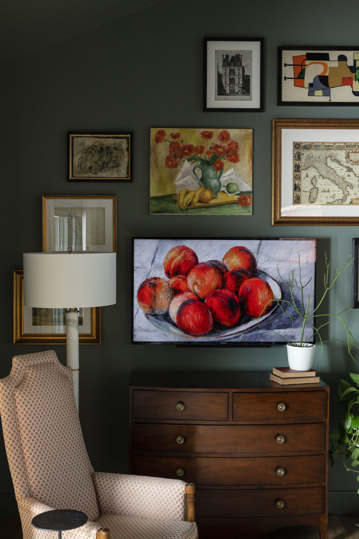 A gallery wall of vintage art surrounding a television displaying a still-life painting, above a mahogany dresser, with a diamond-patterned wingback chair and a marble floor lamp