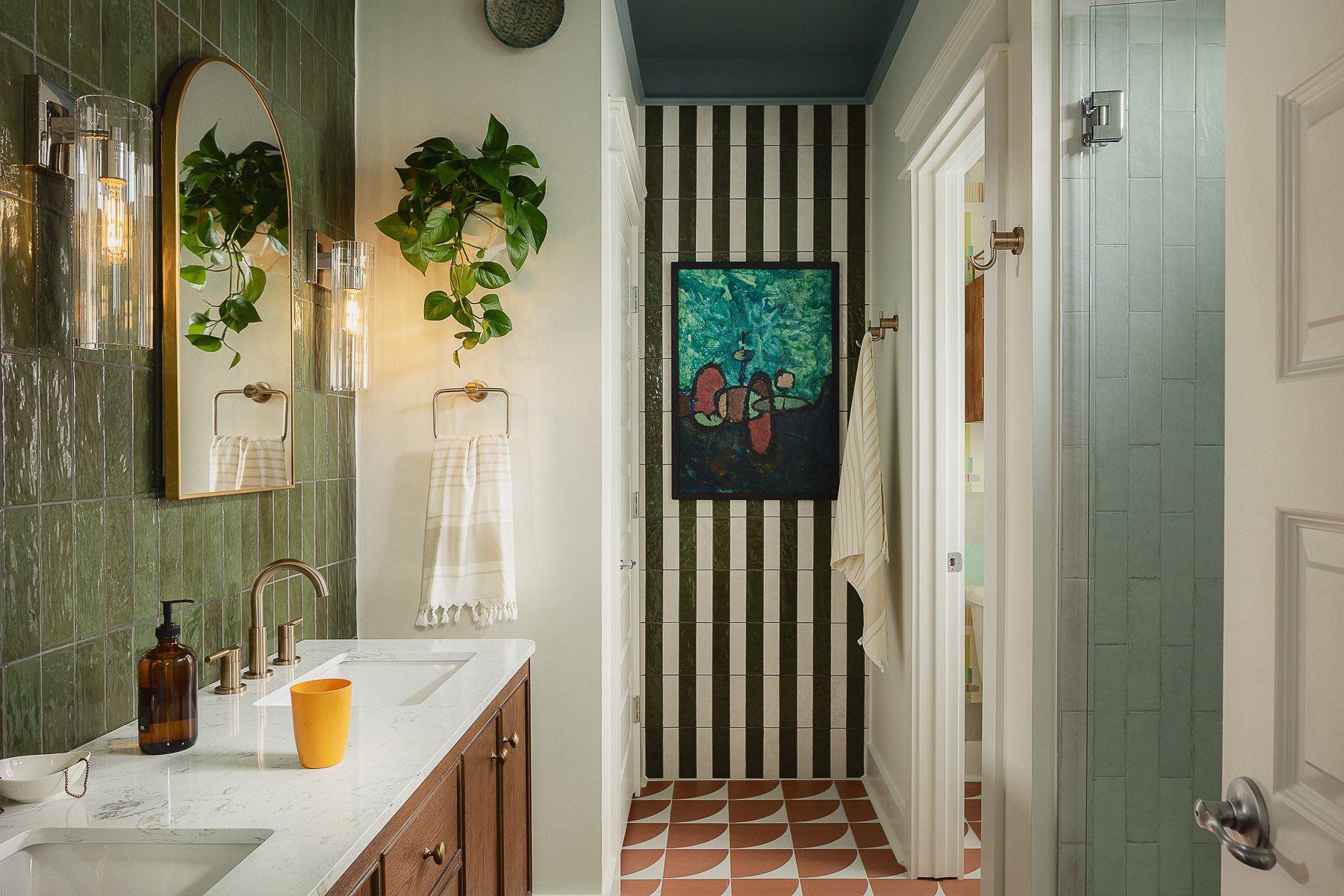 The bathroom: glossy green tile around a brass-framed mirror, a walnut double vanity, a green-and-white striped tile hallway wall with abstract art, and patterned terracotta floor tile