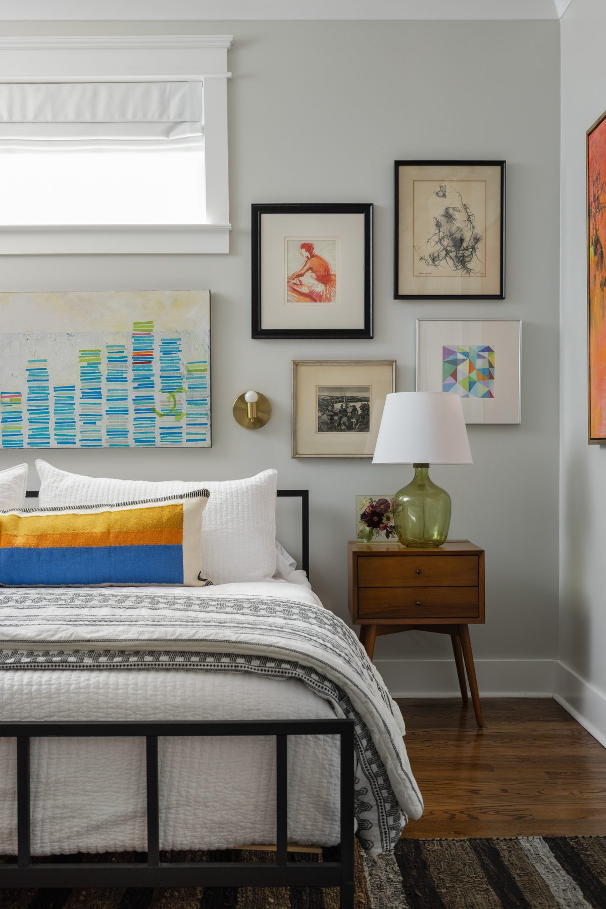 A bright bedroom with a black metal bed, a striped lumbar pillow, a colorful gallery wall, a green glass lamp, and a mid-century nightstand