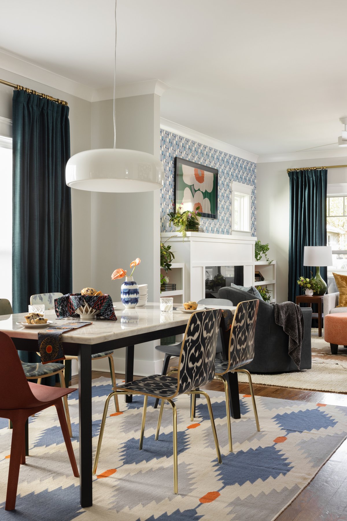 The dining area: a marble-top table with mismatched chairs in rust and ikat prints, a flat-woven rug in blue and orange, and the wallpapered fireplace wall beyond