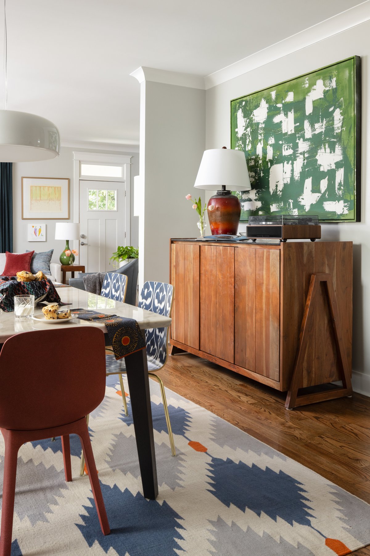 A warm wood sideboard with a record player, a red ceramic lamp, and a large green abstract painting above