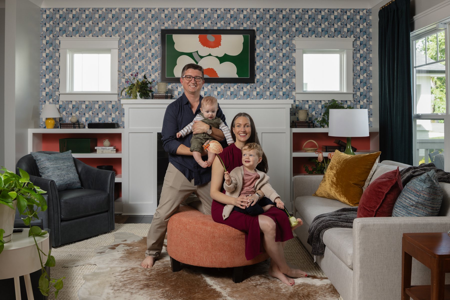 The family of four gathered on a coral ottoman in front of the fireplace, framed by a graphic blue-and-white wallpapered wall with coral-backed built-in shelves