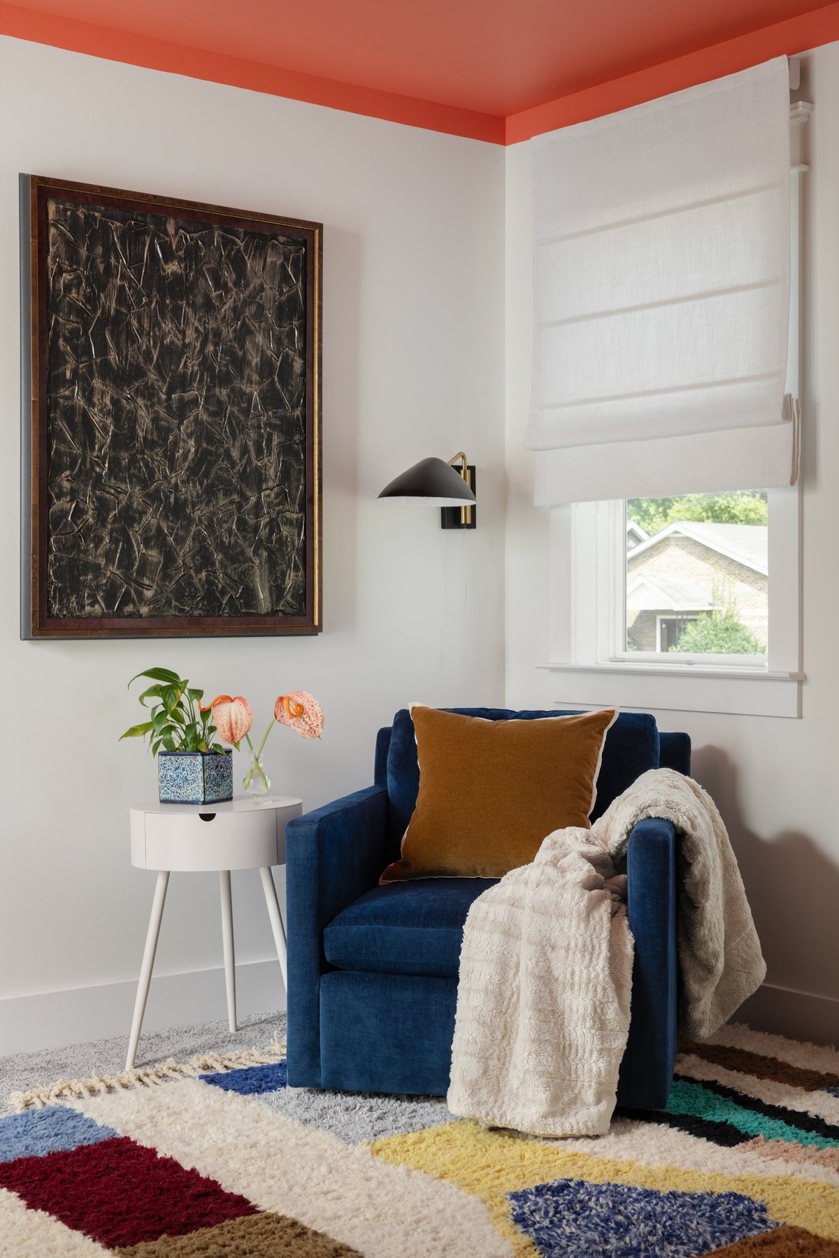 The office reading corner: a navy velvet armchair with a marigold pillow and fur throw under the coral ceiling, beside a textured black painting and a white side table with anthuriums