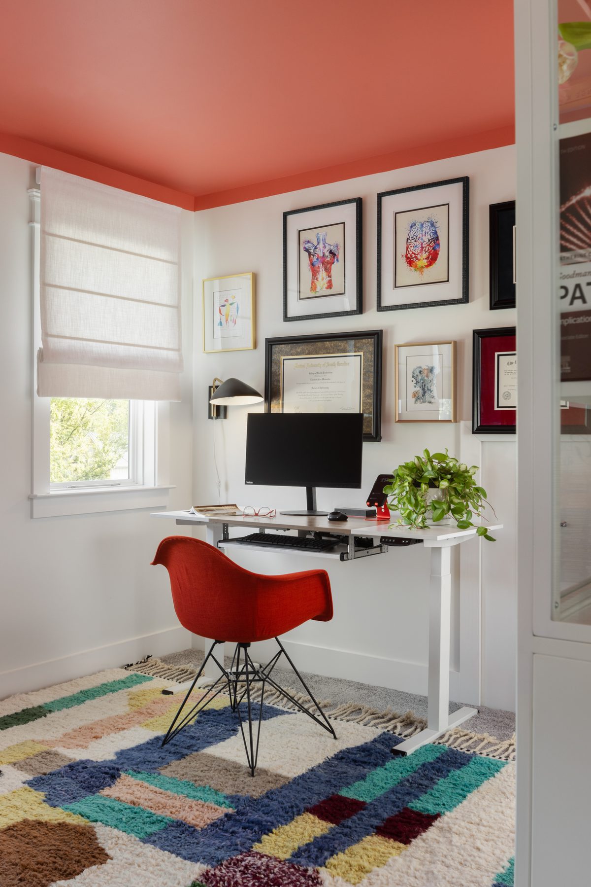 The office: a coral-painted ceiling over a white sit-stand desk, a red Eames-style chair, a gallery wall of framed diplomas and anatomy art, and a color-blocked shag rug