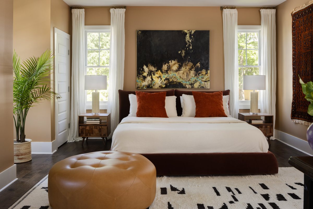 A bedroom with warm tan walls, a velvet bed with rust pillows, a large abstract painting, and a leather ottoman