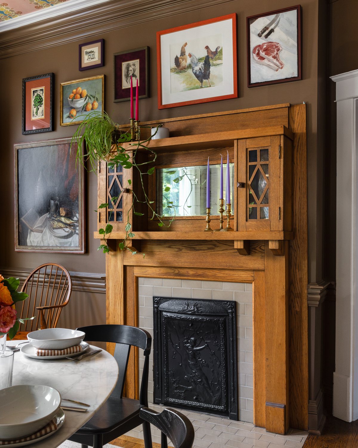 A Craftsman oak mantel with a gallery wall, brass candlesticks, and a wallpapered ceiling in a dining room