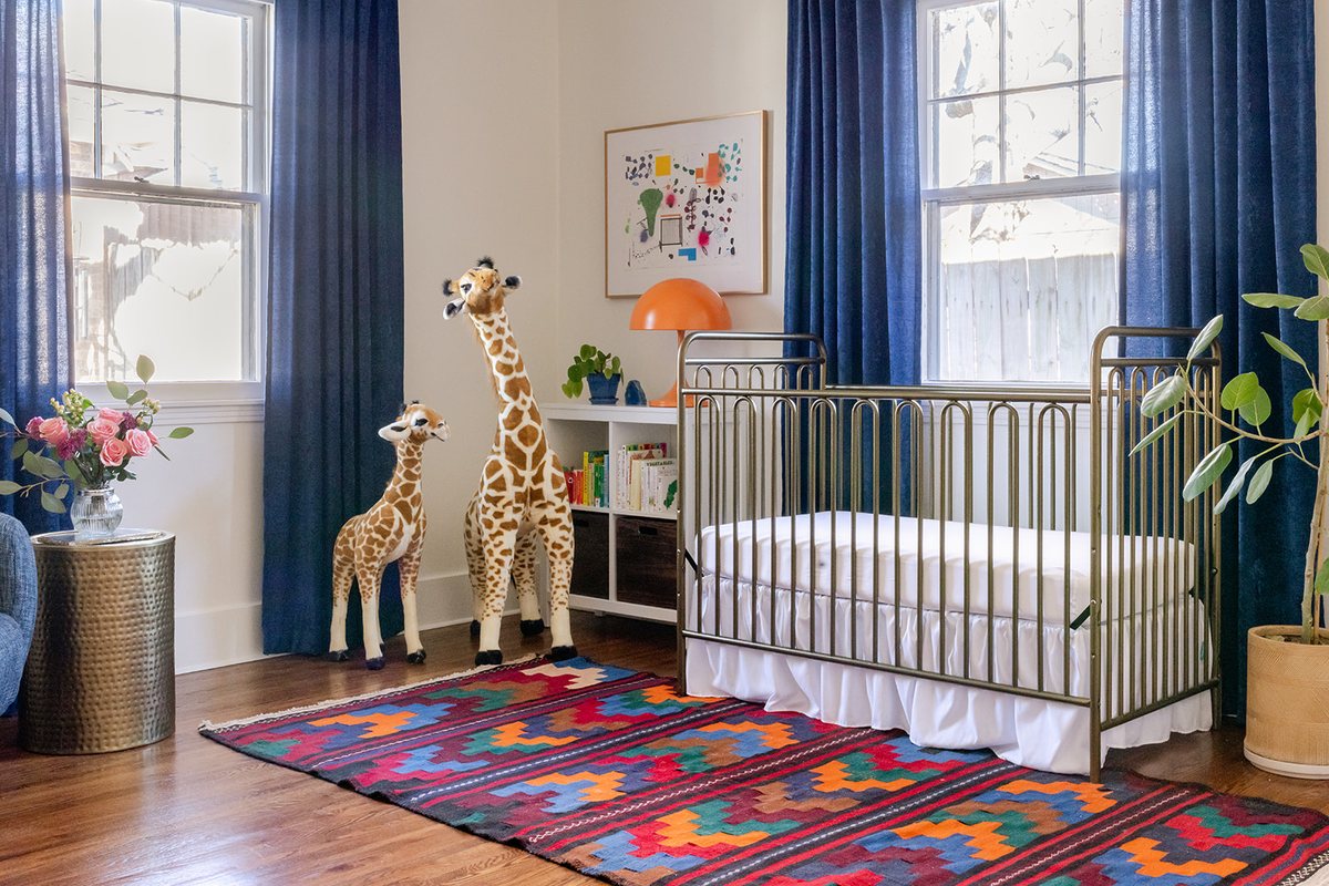 A nursery with a brass crib, a colorful vintage kilim, navy velvet drapes, and a pair of plush giraffes