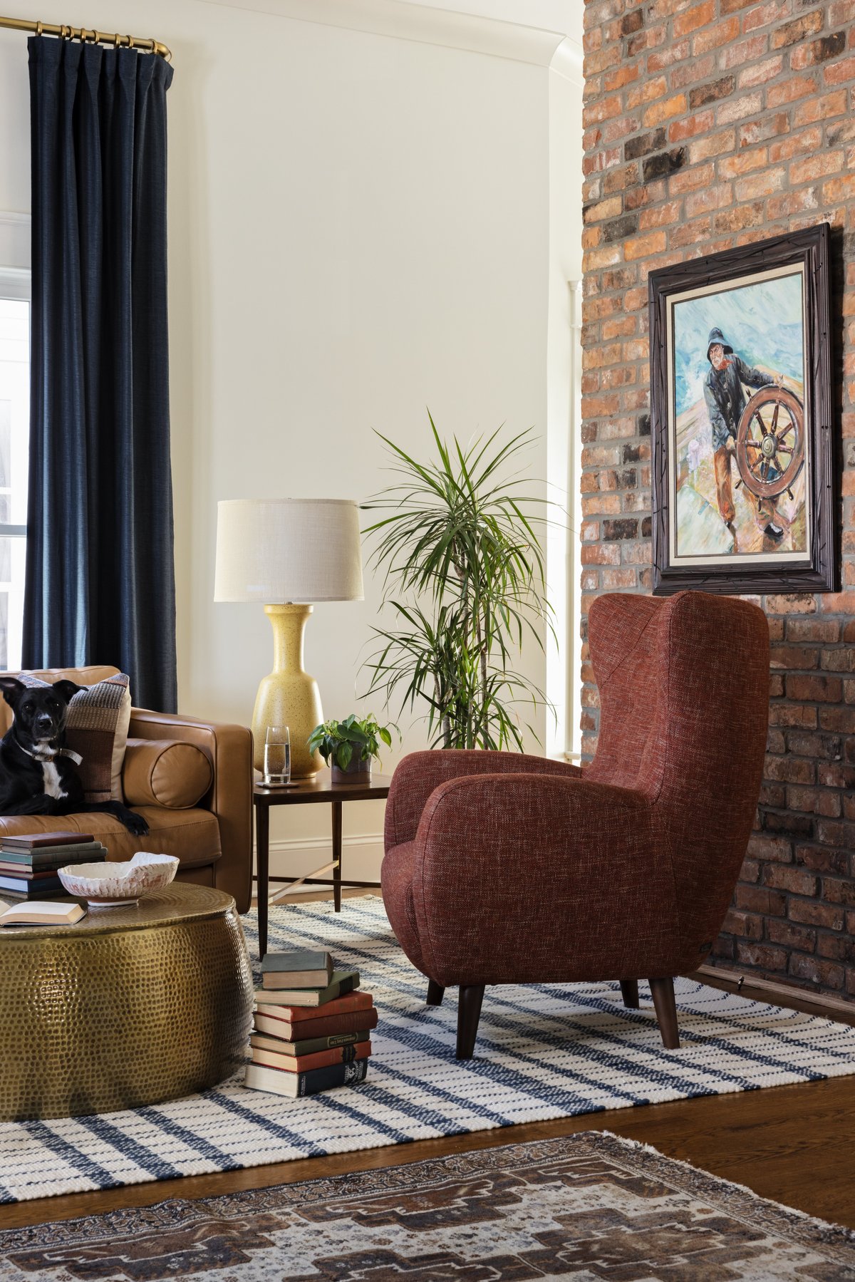 The other rust wingback against the exposed brick fireplace with a vintage sea-captain painting, and the family&rsquo;s black dog lounging on the caramel leather sofa
