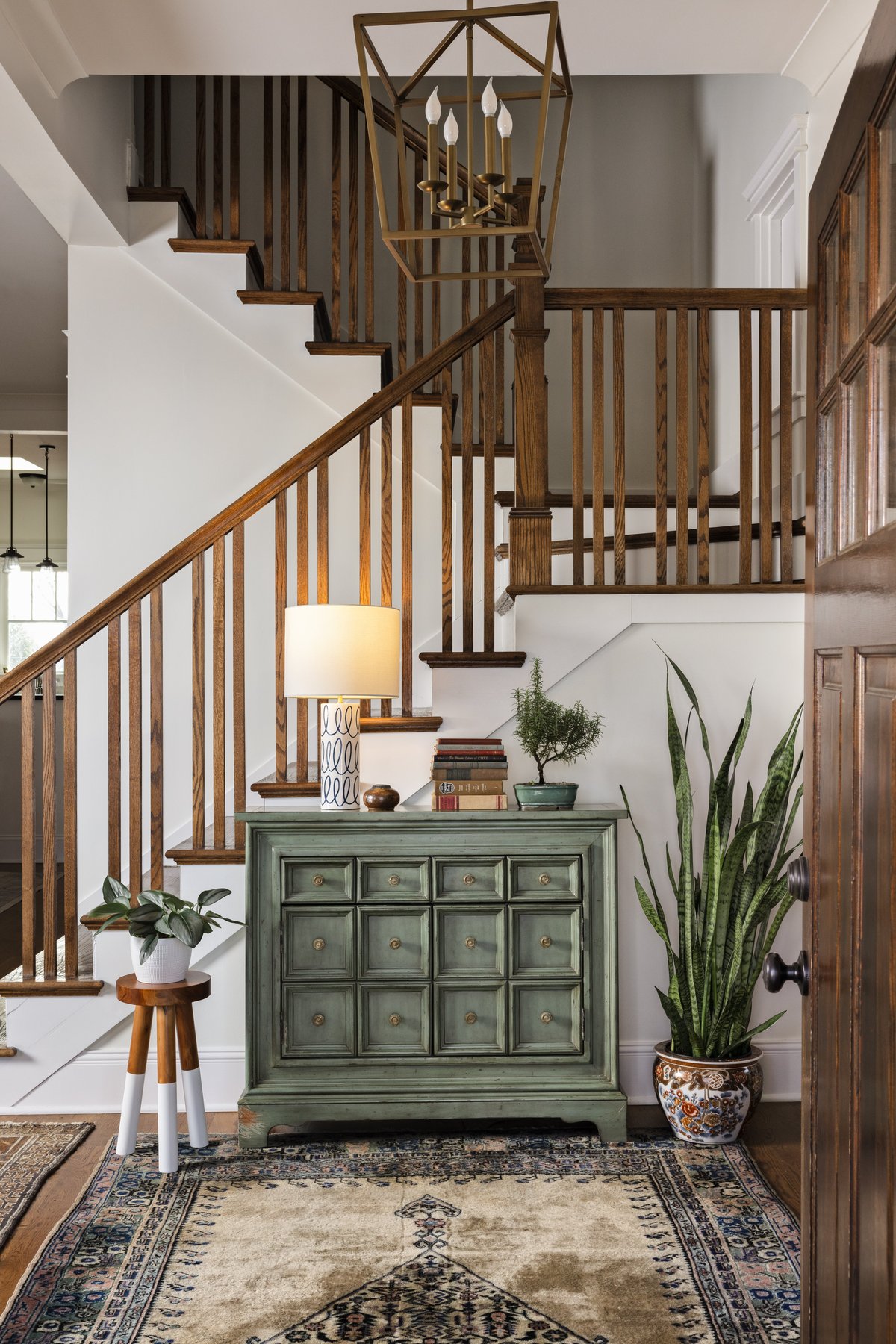 The entry: a green apothecary-style chest under the staircase with a squiggle-patterned lamp, stacked antique books, a bonsai, and a snake plant in a chinoiserie planter on a vintage rug