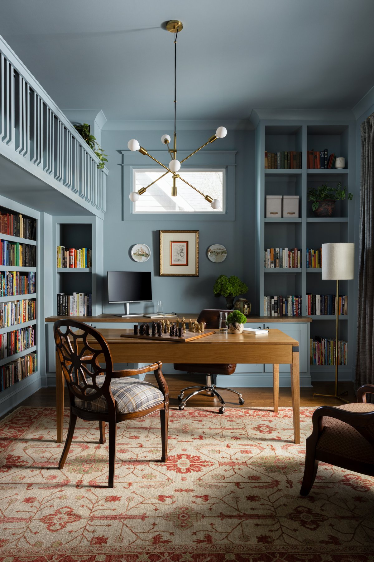 The family library: slate-blue built-in bookshelves on both walls, a wood desk with a chess set mid-game, a plaid spider-back chair, a brass chandelier, and the loft railing above