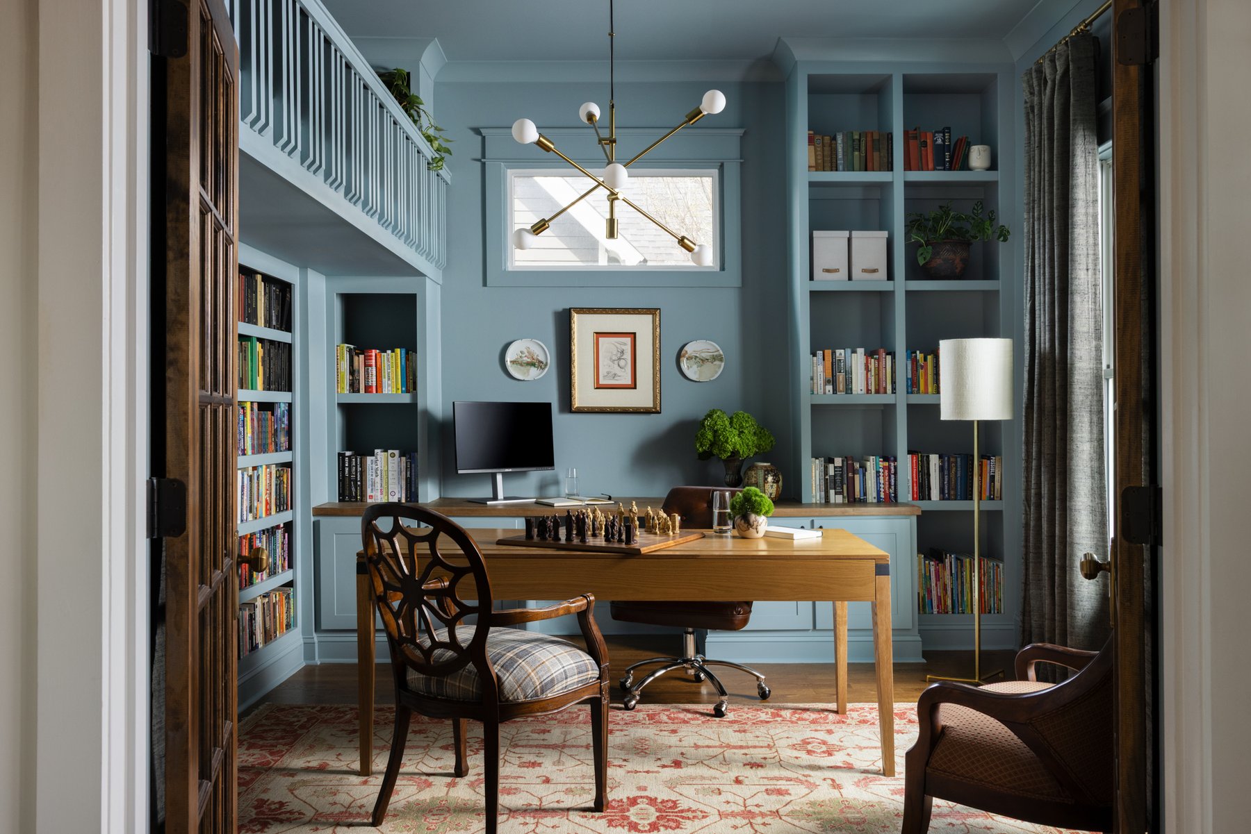 The family library: slate-blue walls and built-in bookshelves on both sides, a wood desk with a chessboard mid-game, a reupholstered spider-back chair in plaid, a brass mobile chandelier, and the loft railing above