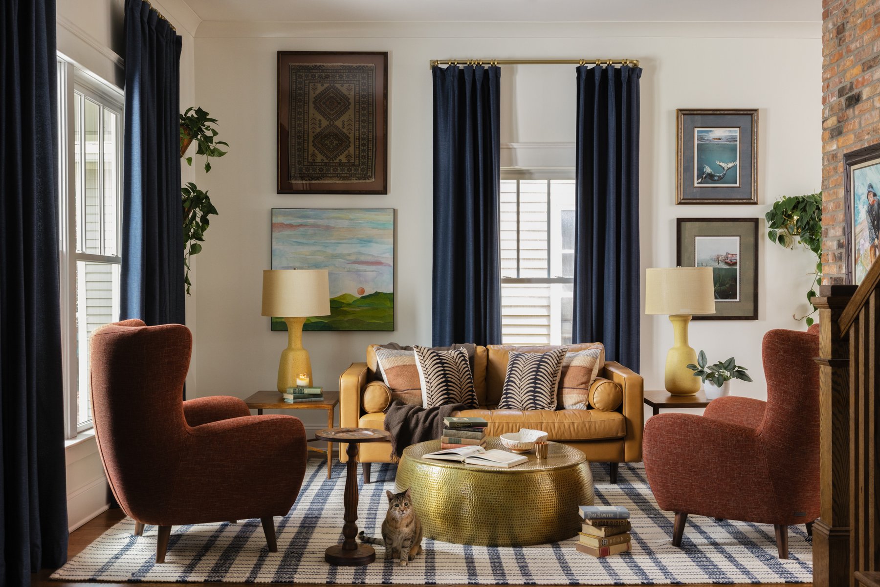 The living room: a mustard leather sofa with patterned pillows flanked by two rust wingback chairs, navy drapes, a hammered brass coffee table, a framed vintage rug on the wall, and the family&rsquo;s tabby cat on the plaid rug