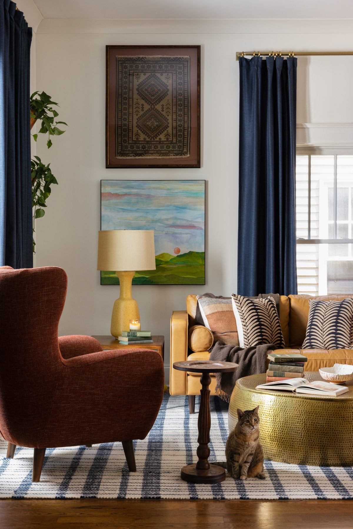 A rust wingback chair beside the mustard leather sofa, a torn-paper landscape painting and framed vintage rug on the wall, and the tabby cat under the brass coffee table