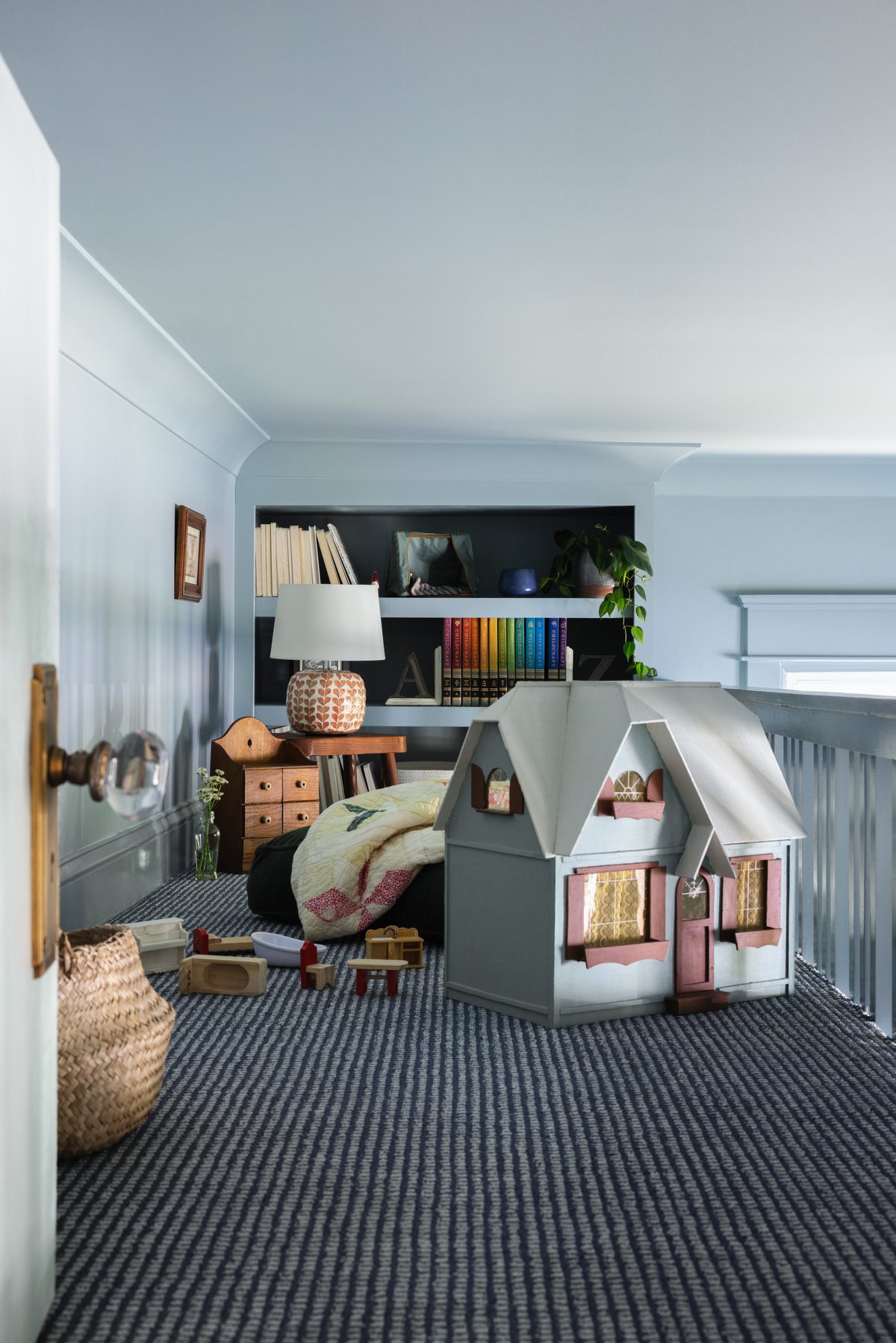 Inside the loft kids&rsquo; space: a big dollhouse and wooden toy furniture on striped navy carpet, a quilt on a floor cushion, and a built-in bookcase with a rainbow of Childcraft volumes at the back