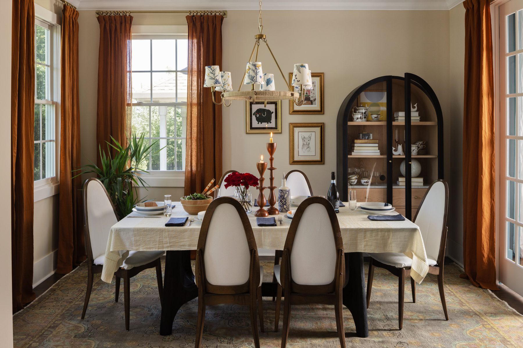The dining room: a table set beneath a brass chandelier, oval-back chairs, rust drapes, a black arched display cabinet, and candlesticks on a white tablecloth
