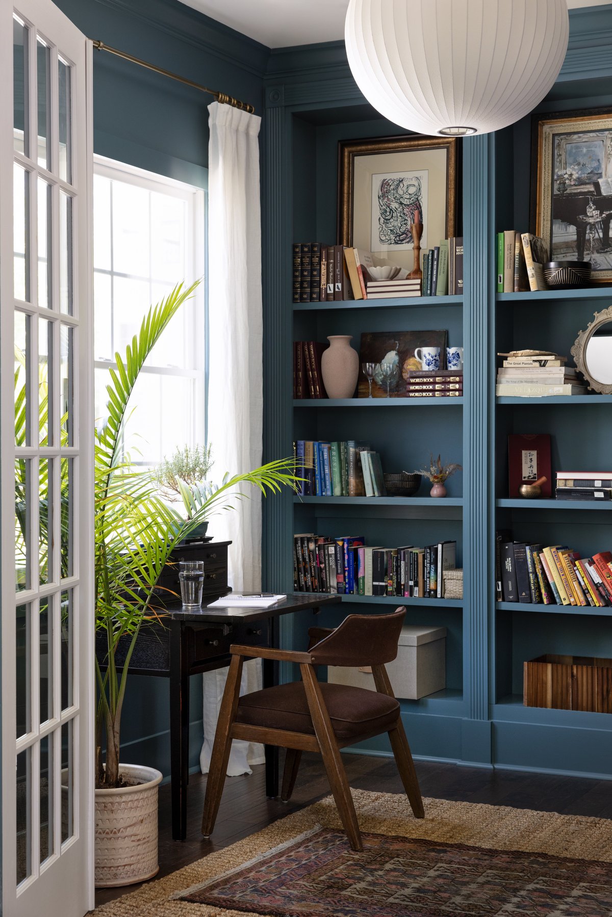 Her office through the French doors: a black writing desk with a vintage chair by the window, a palm, and slate-blue built-in shelves styled with books and art