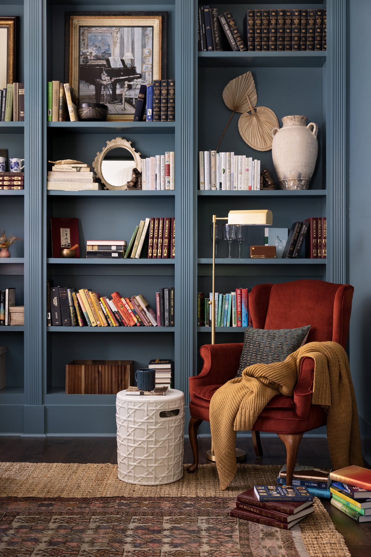 The office built-ins in slate blue with collected books, a vintage mirror, dried palms, and a rust wingback chair with a mustard throw beside a brass floor lamp