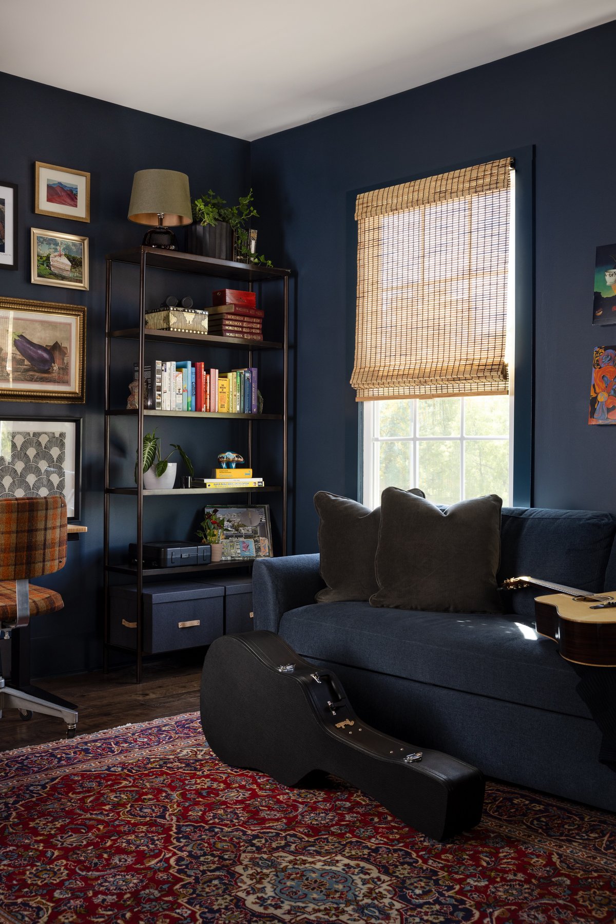His office in deep navy: a gallery wall of collected art over the desk with a plaid vintage task chair, open shelving with color-blocked books, and a navy sofa with a guitar case