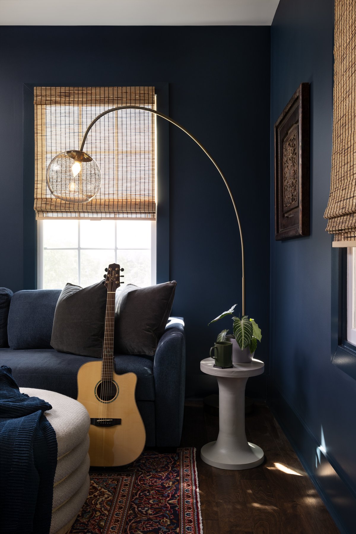 The other corner of his office: a brass arc lamp over the navy sofa with an acoustic guitar, bamboo shades, and a carved wood panel on the wall