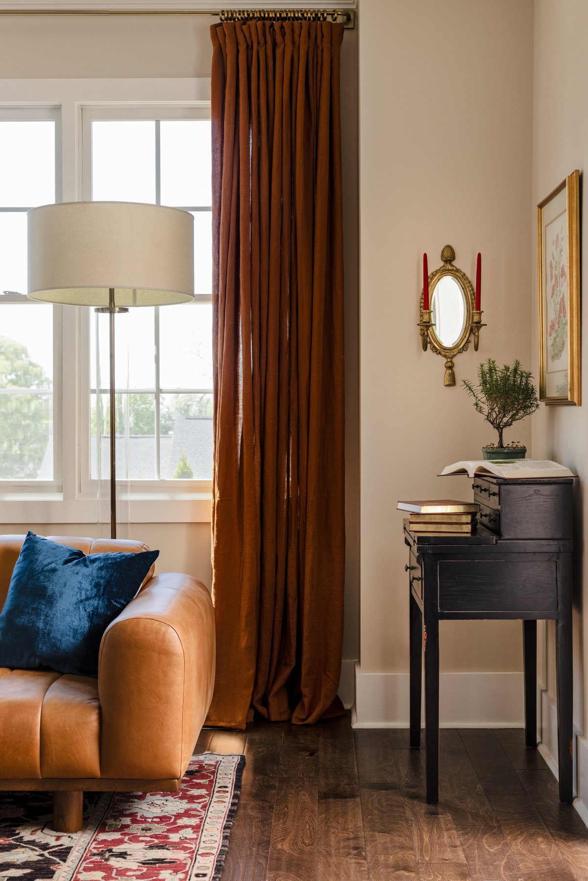A corner of the living room: the caramel leather sofa with a navy velvet pillow, rust drapes, and a black secretary desk under a gilt candle sconce with red tapers