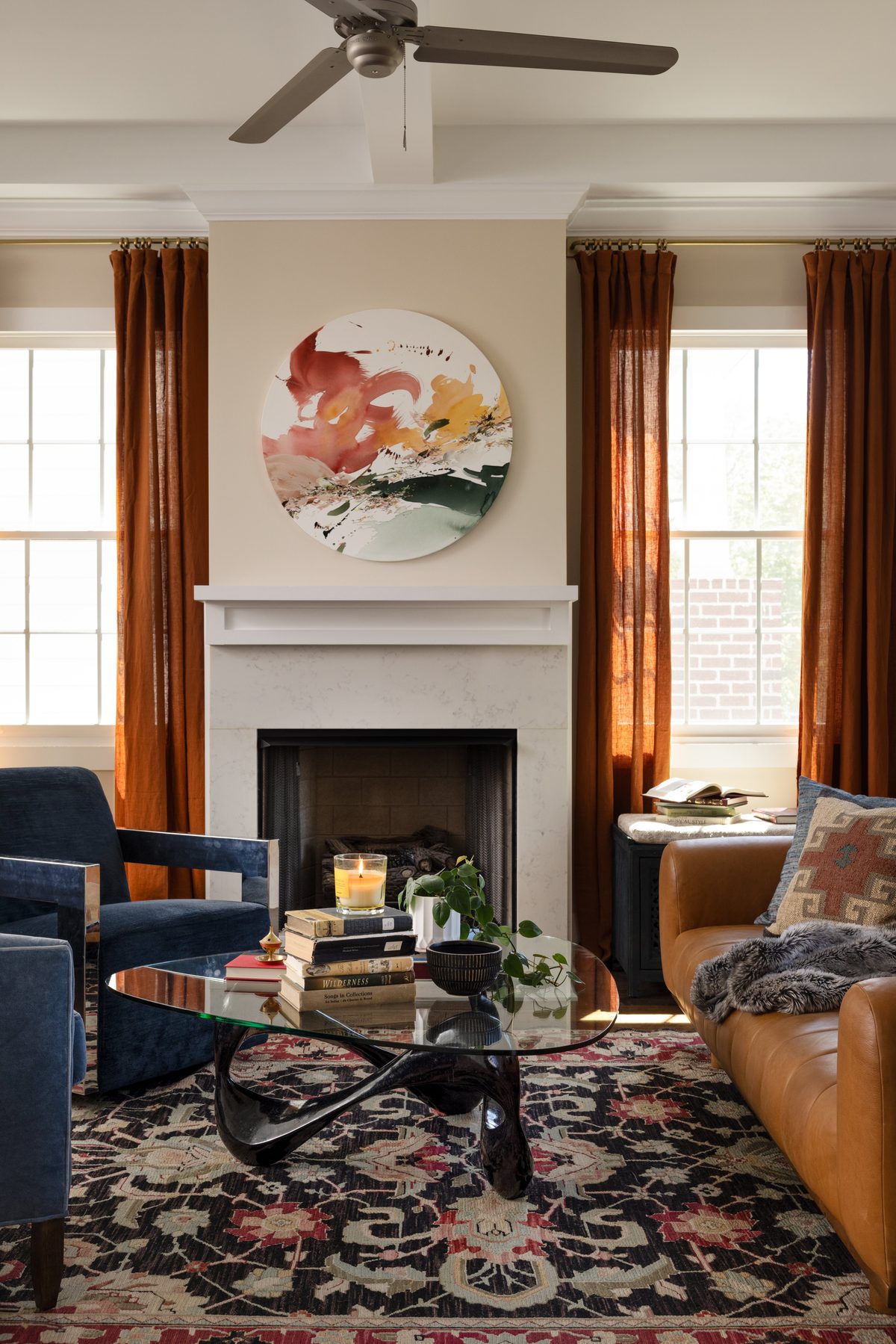 The living room: navy velvet chairs and a caramel leather sofa around a glass Noguchi-style coffee table, rust linen drapes, a floral vintage rug, and a round abstract artwork over the marble fireplace