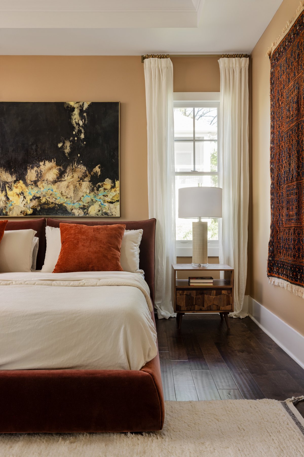 The primary bedroom: a rust velvet bed with terracotta pillows beneath a large black-and-gold abstract painting, with a vintage textile hung on the wall beside it