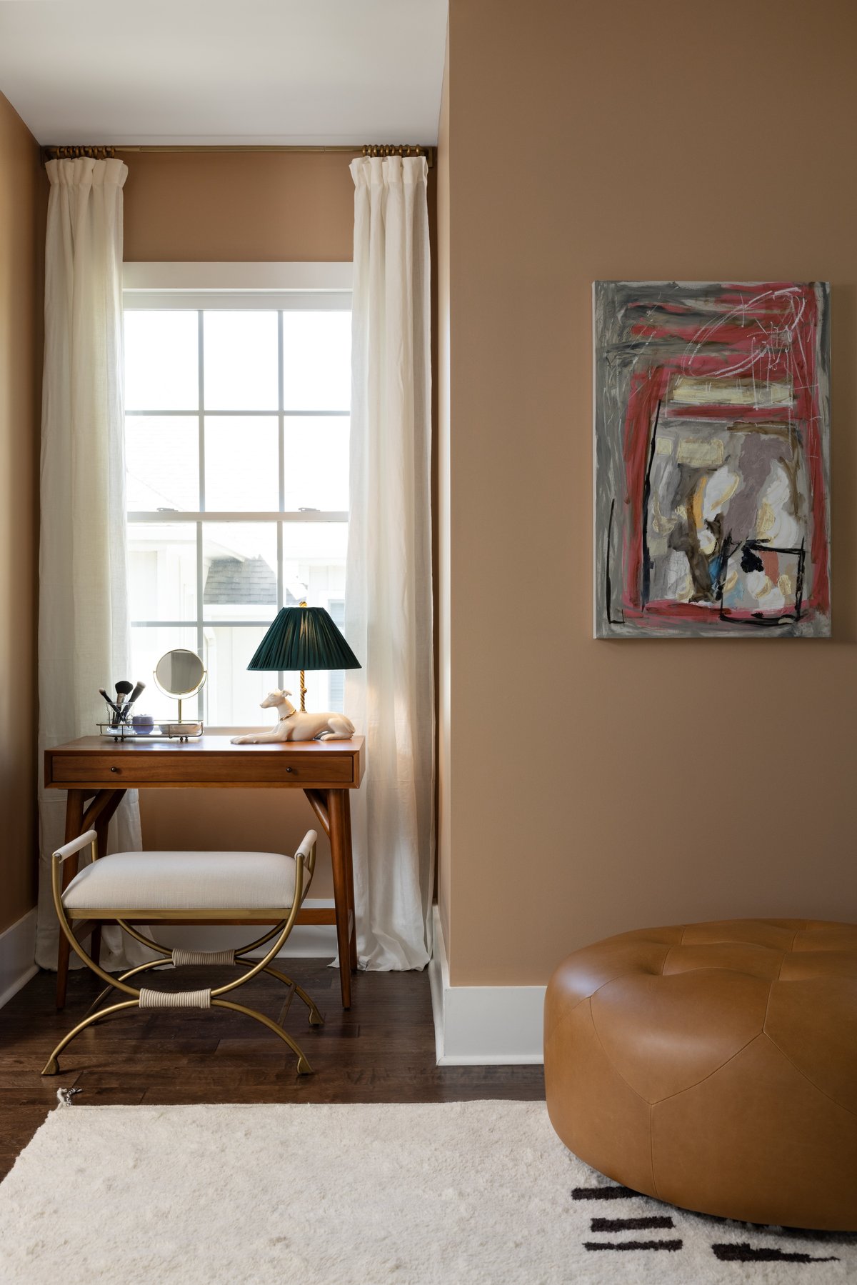 A vanity corner in the primary bedroom: a mid-century desk with a greyhound lamp under a green pleated shade, a brass X-bench, and an abstract painting on the caramel wall