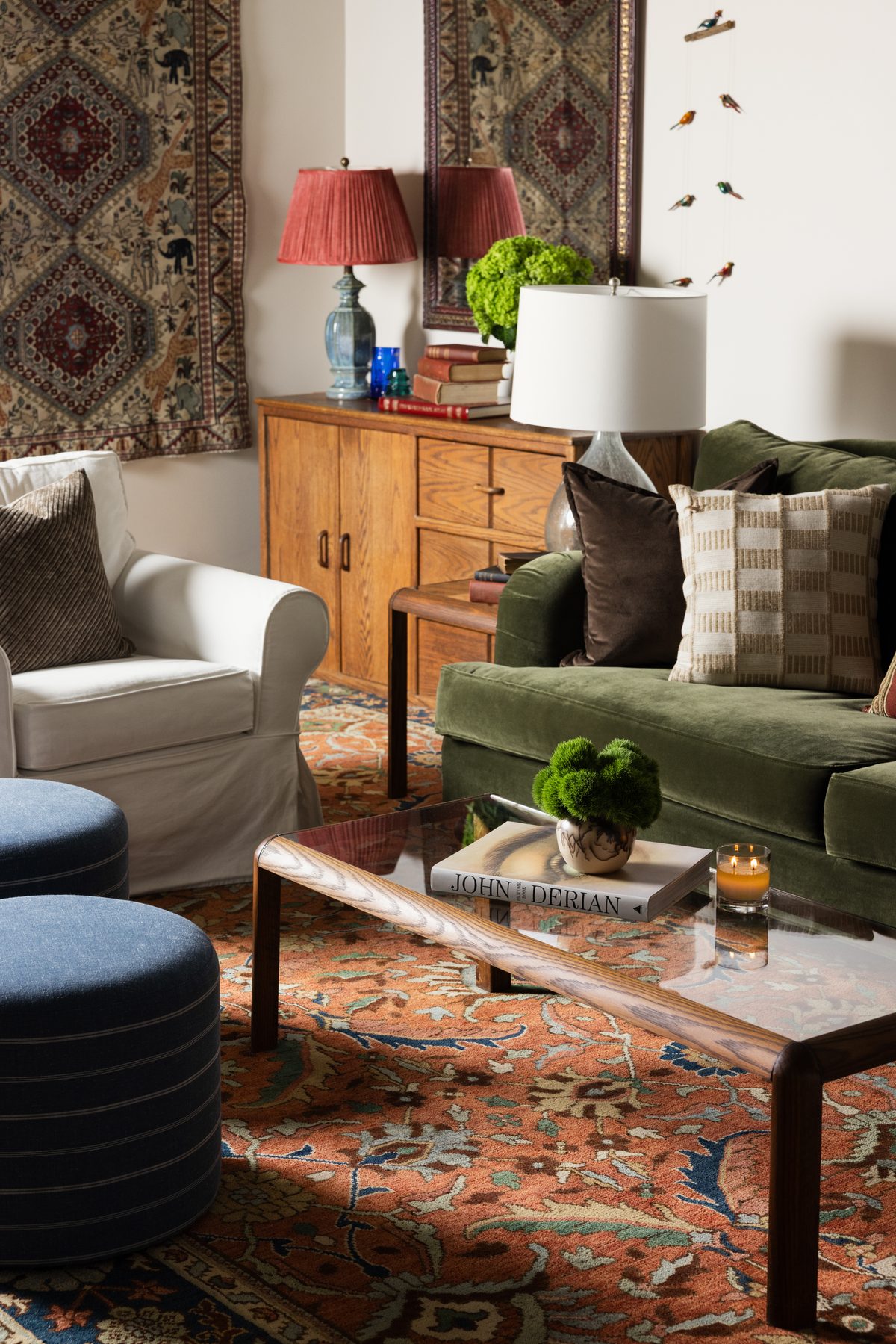 The green velvet sofa beside an oak credenza with a red-shaded lamp, stacked books, and hung textile rugs, with a glass coffee table in the foreground
