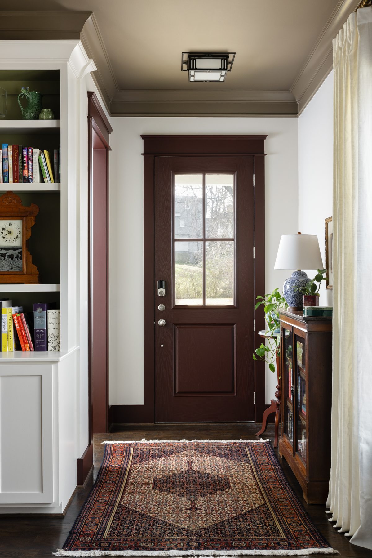 The entry with the front door and trim painted a deep maroon, a taupe ceiling, built-in bookshelves, and an antique glass-front cabinet on a Persian runner