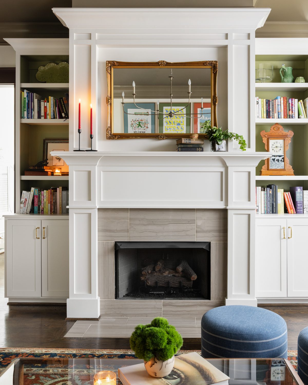 The white fireplace and built-ins with olive-backed shelves, a gilt mirror over the mantle, red taper candles, and a striped blue pouf